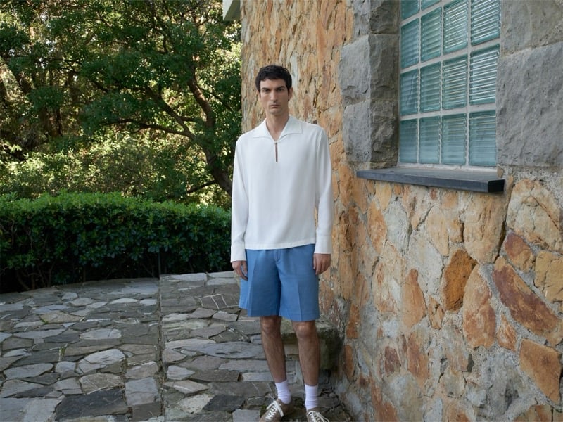 Luca Lemaire stands in a white tunic and blue shorts against a stone wall surrounded by greenery