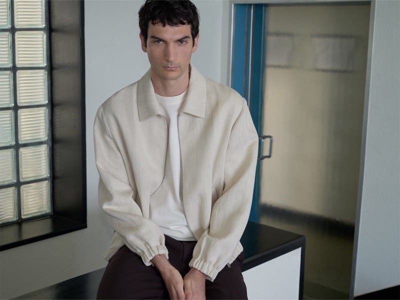 Luca Lemaire sits with his hands clasped in a cream jacket over a white tee and dark trousers near a glass block window