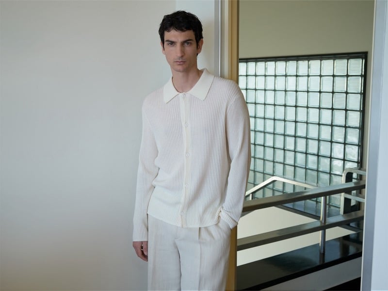 Luca Lemaire wears a cream ribbed knit cardigan and matching trousers, leaning against a wall near a glass block window