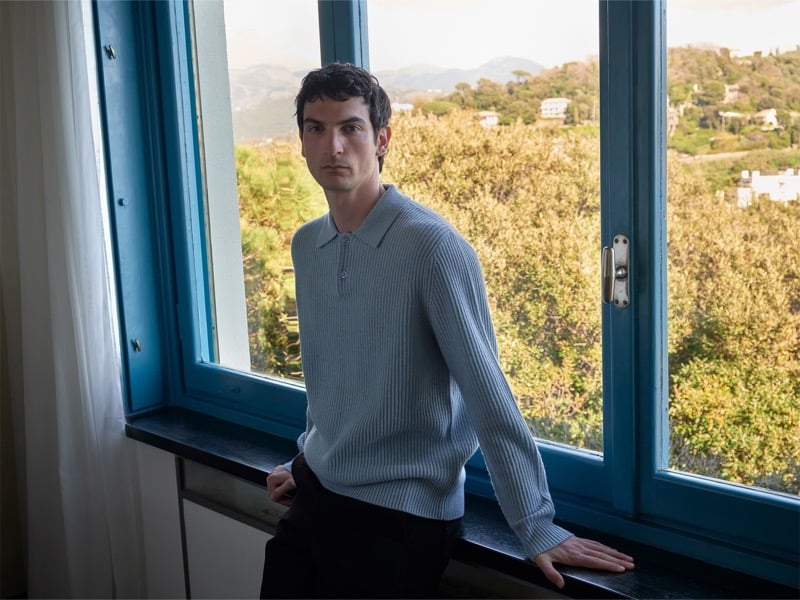 Luca Lemaire leans against a blue window frame in a pale blue-gray ribbed polo, hills and greenery visible outside