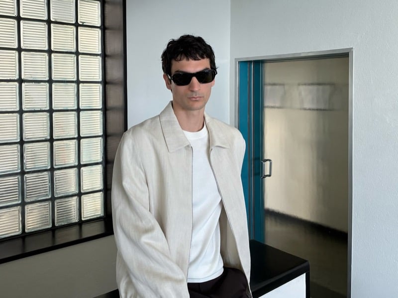 Luca Lemaire sits on a black bench in sunglasses and a cream jacket over a white tee and dark trousers