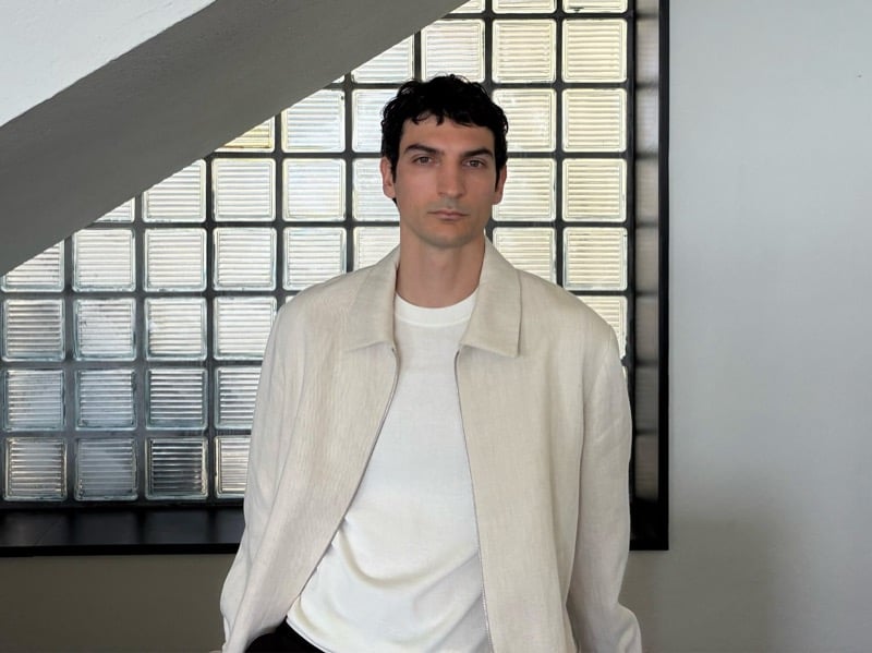 Luca Lemaire stands beneath a staircase in a cream jacket over a white tee, a glass block window behind him