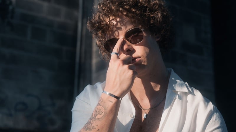 A curly-haired man in sunglasses and a white linen shirt sits wearing a silver cuff bracelet