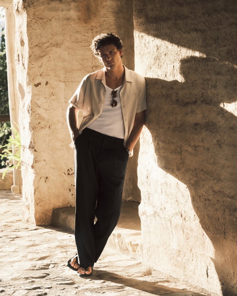 Simon Nessman wears an open cream camp-collar shirt over a white tank with dark pleated trousers leaning against a sunlit stone archway for Calvin Klein