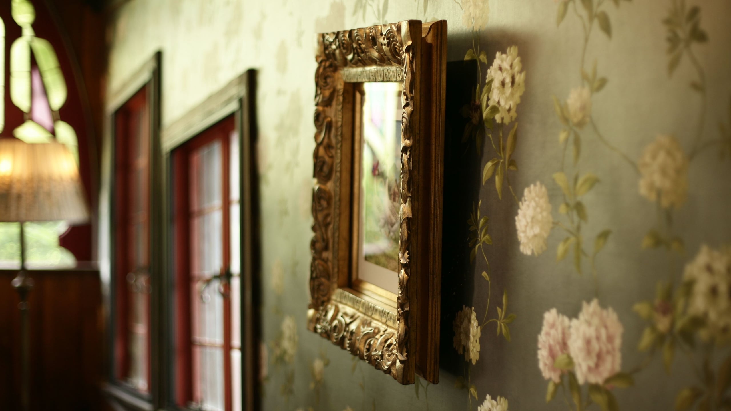 A gilt-framed painting hangs on a wall covered in small pale floral wallpaper