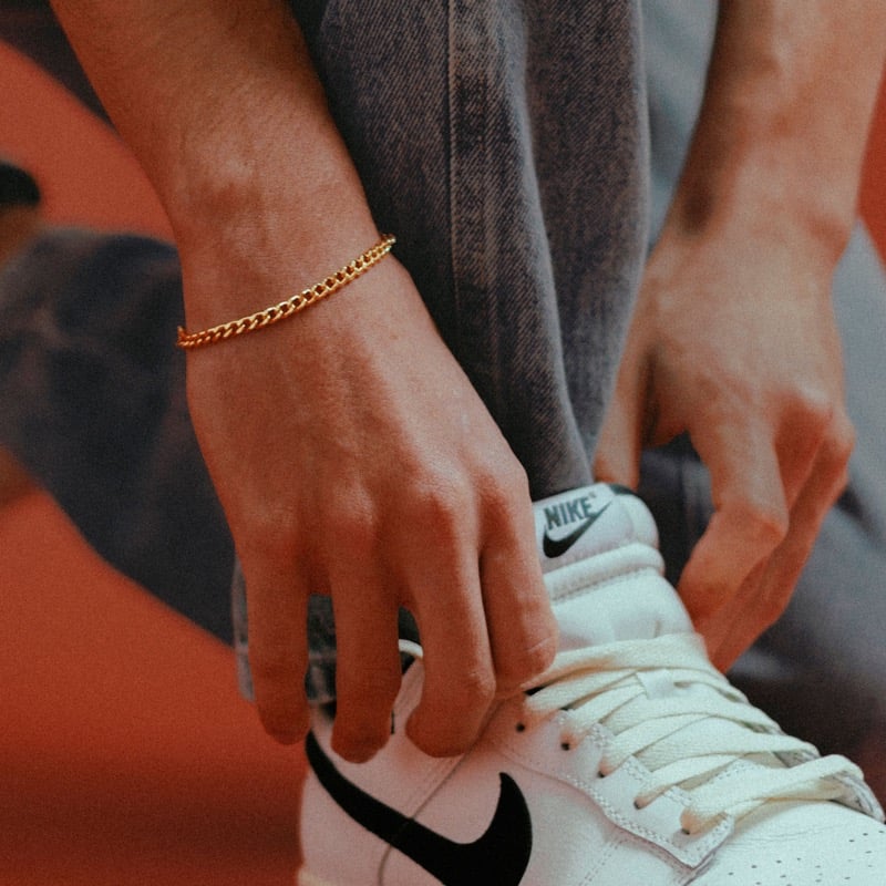 A man's wrist wearing a gold curb chain bracelet while tying a white sneaker