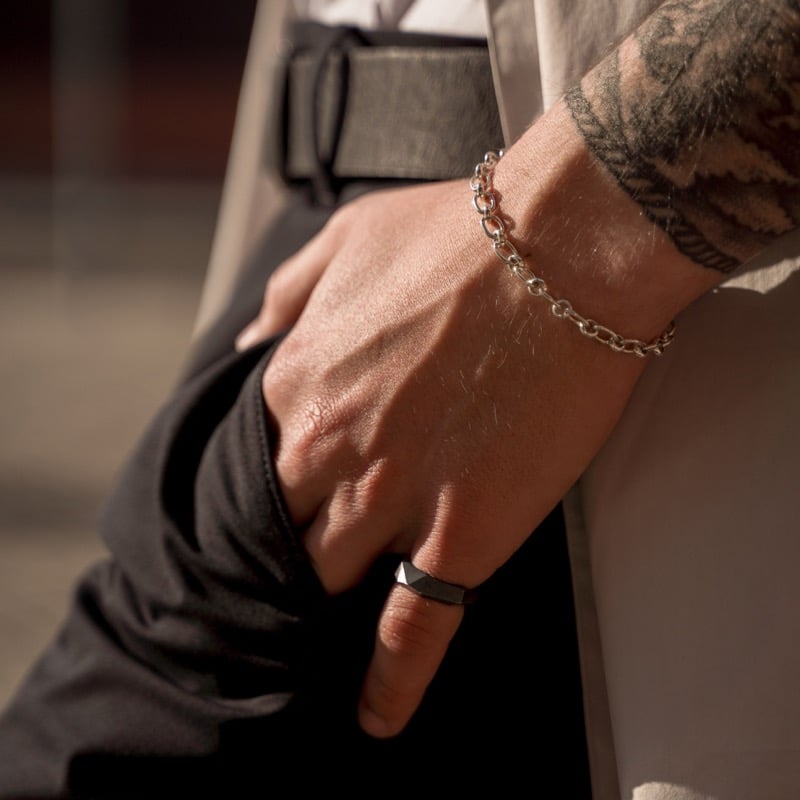 A man's tattooed wrist wearing a stainless steel chain bracelet and a black ring