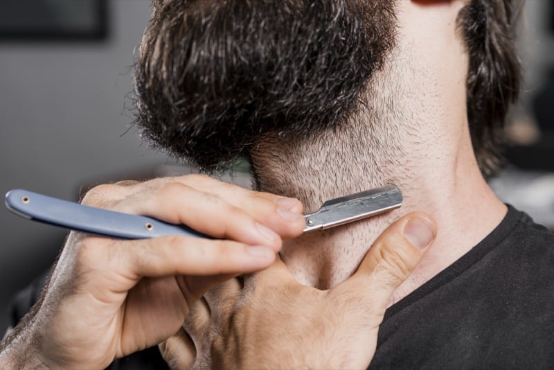 A straight razor drawn along the side of a bearded man's neck in close-up