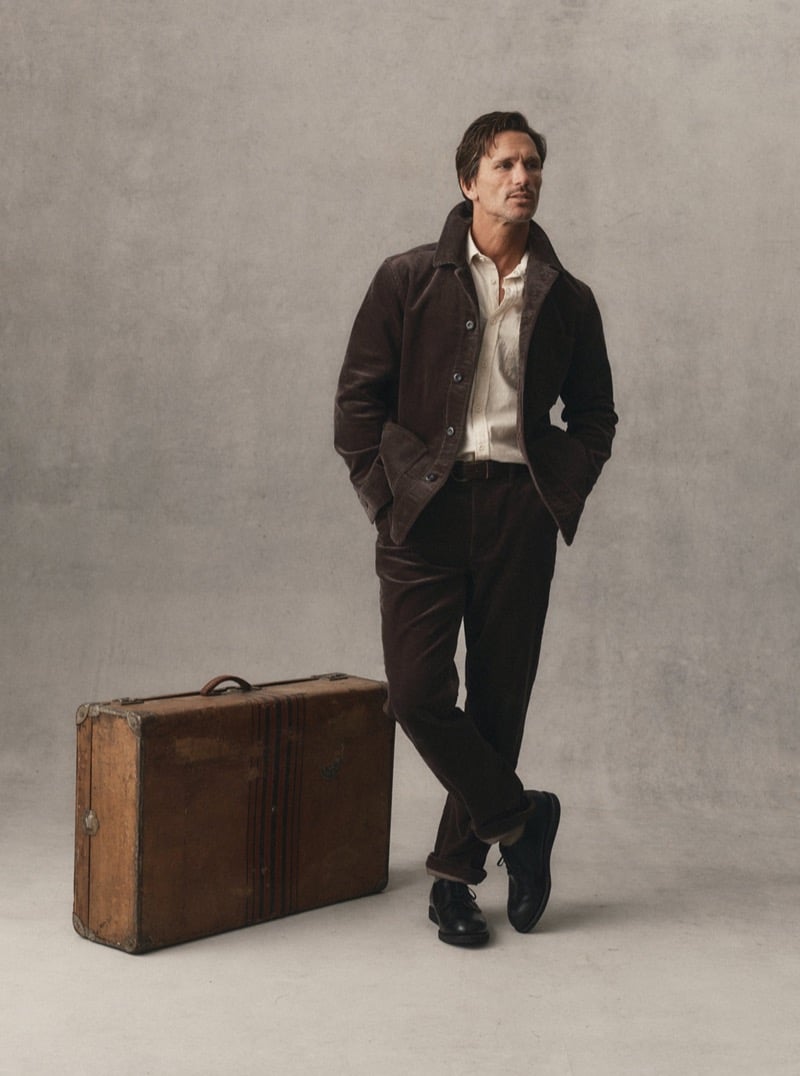 Man in a dark brown corduroy jacket and trousers with a light button-up shirt leaning against a vintage leather suitcase, for Taylor Stitch Fall 2026
