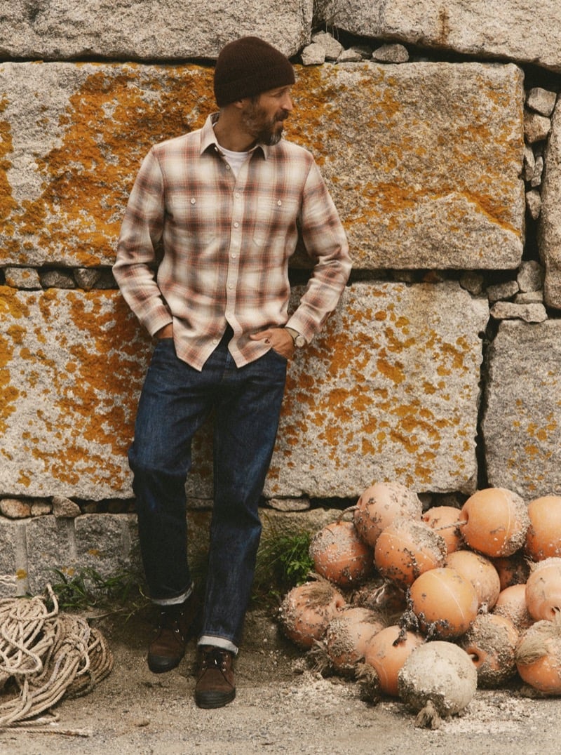 Man in a plaid flannel shirt, knit beanie, and dark jeans leaning against a stone wall beside buoys and coiled rope, for Taylor Stitch Fall 2026