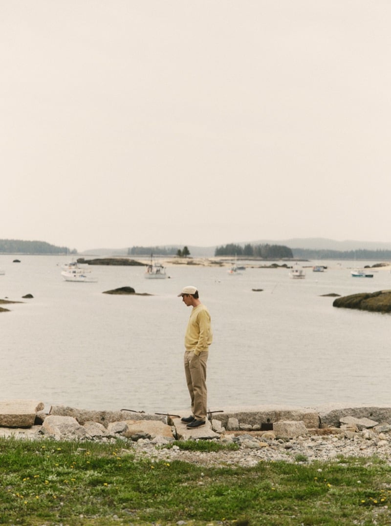 Man in a yellow sweater and khaki pants standing on a rocky shoreline overlooking a harbor full of fishing boats, for Taylor Stitch Fall 2026