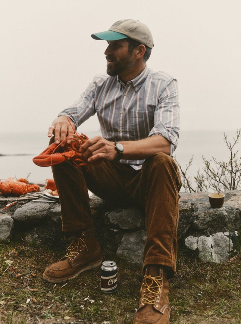 Man in a striped shirt and brown corduroy pants sitting on a stone wall by the ocean eating a lobster, for Taylor Stitch Fall 2026