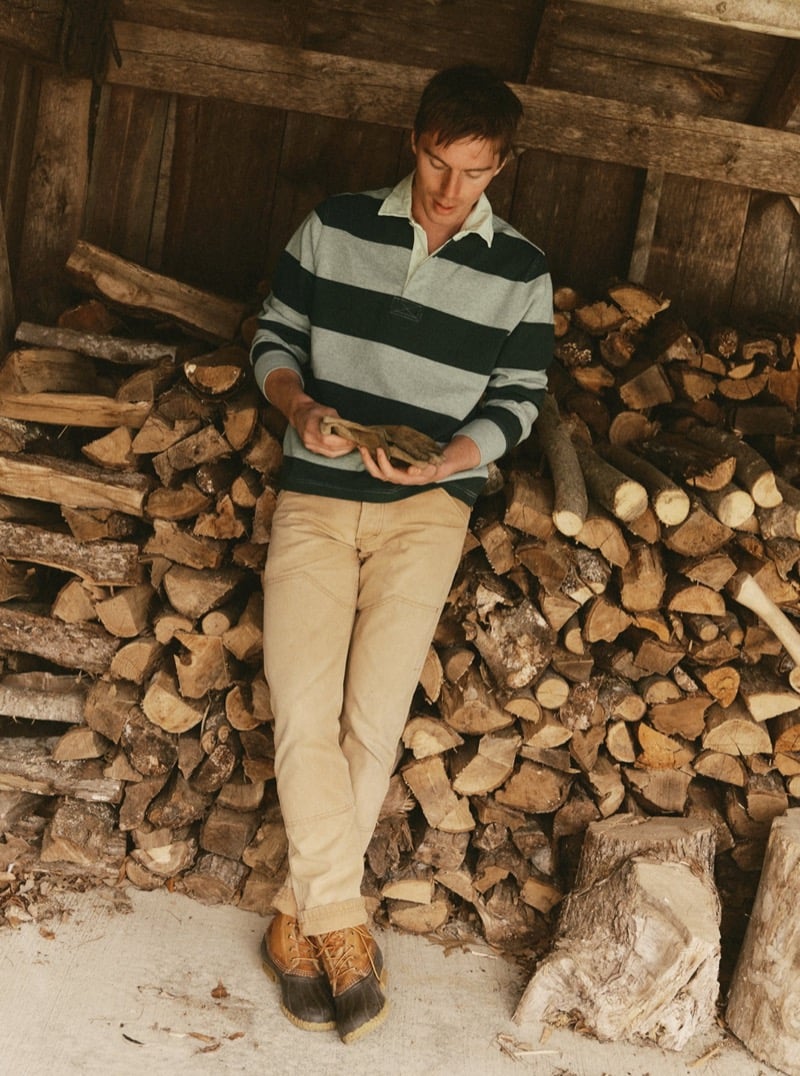 Man in a striped rugby polo and tan pants leaning against a stacked woodpile, for Taylor Stitch Fall 2026