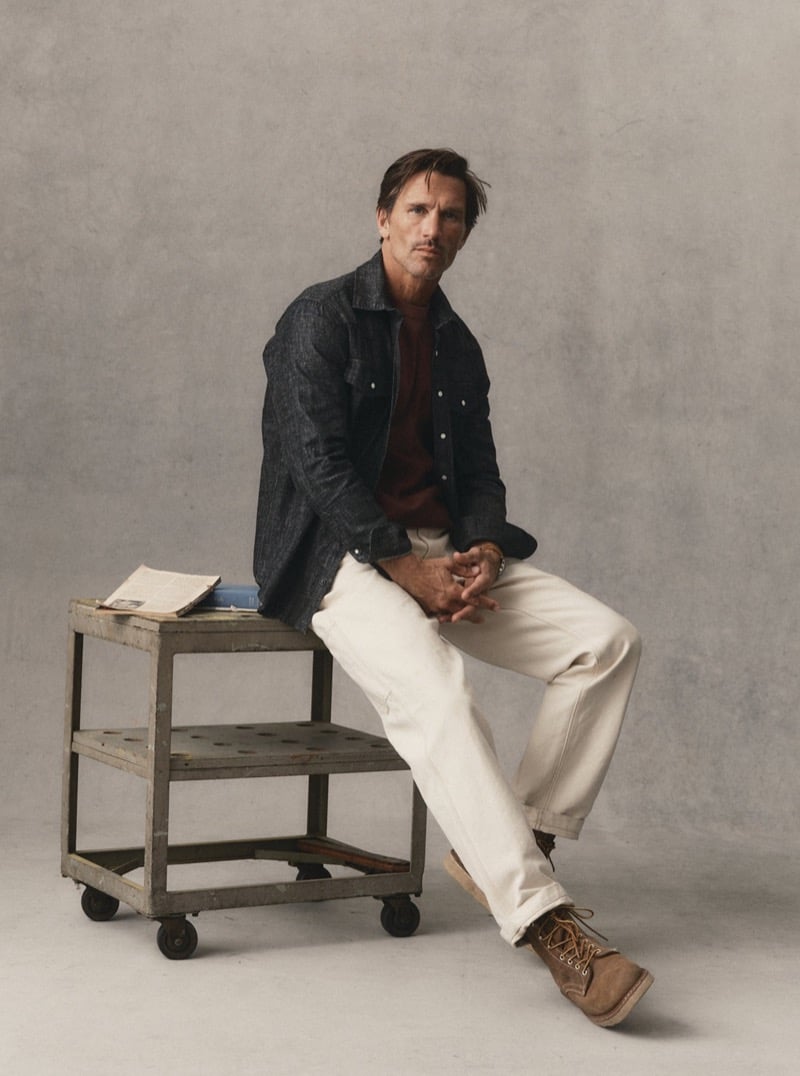 Man in a dark denim overshirt and light trousers sitting on a rolling cart in a studio, for Taylor Stitch Fall 2026