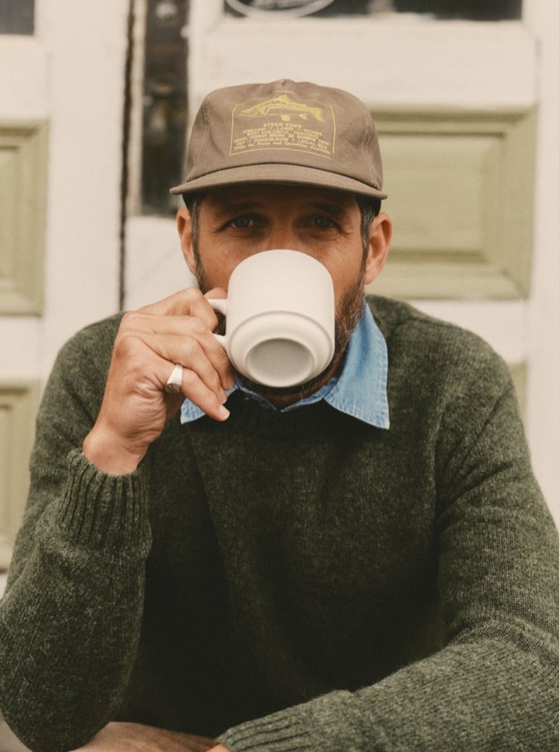 Man in a green sweater and chambray shirt drinking coffee from a mug on a porch, for Taylor Stitch Fall 2026