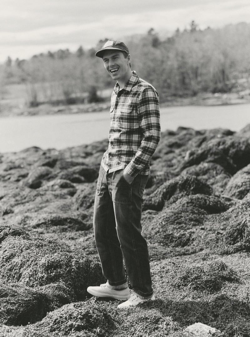 Black-and-white image of a man in a plaid flannel shirt and jeans laughing on seaweed-covered rocks, for Taylor Stitch Fall 2026