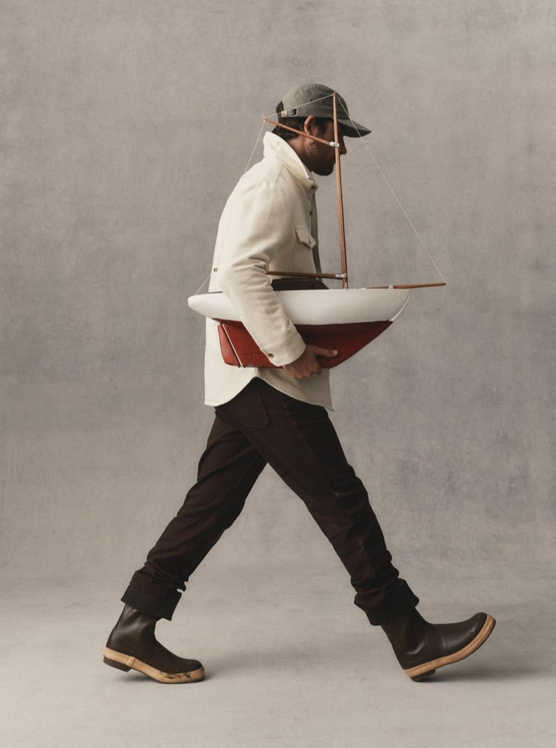 Man in a light wool jacket and dark brown pants carrying a model sailboat in a studio, for Taylor Stitch Fall 2026