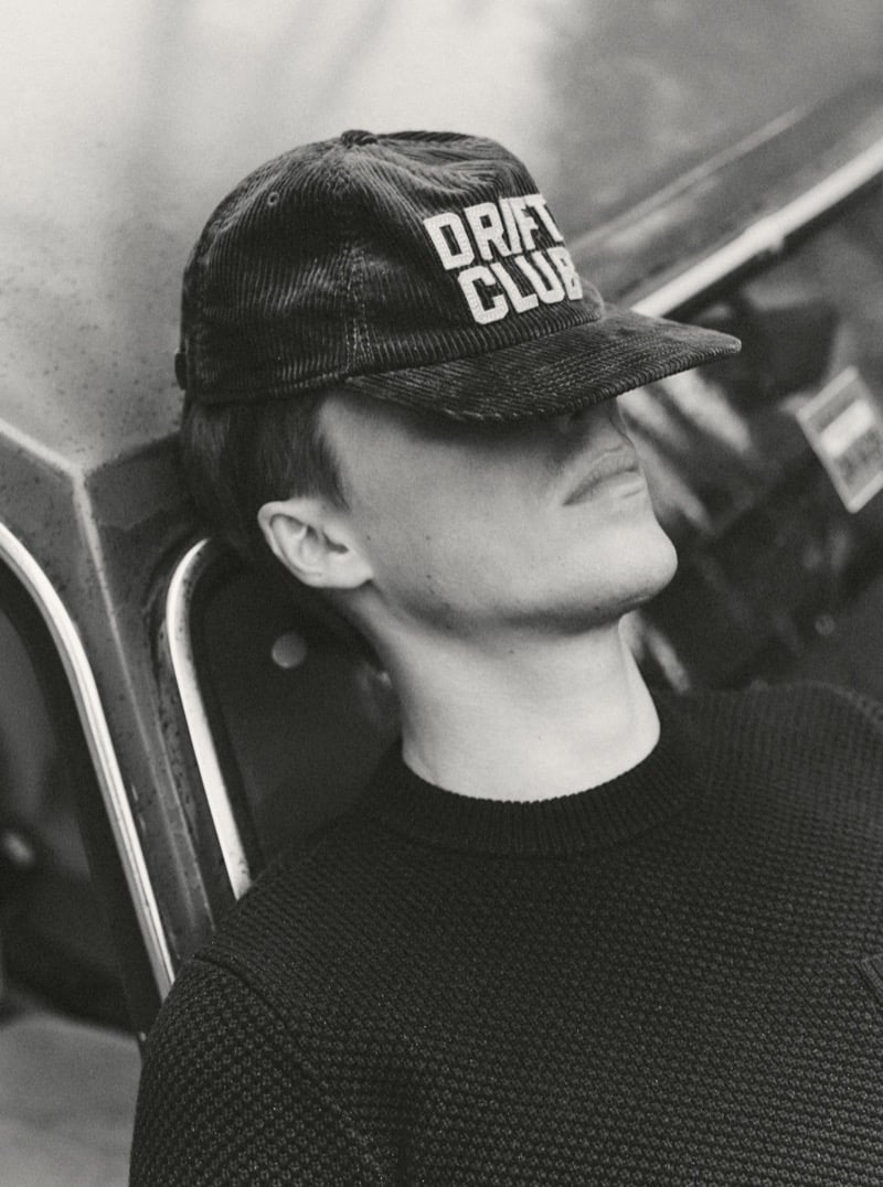 Black-and-white close-up of a man in a corduroy cap and textured sweater leaning against a vintage car, for Taylor Stitch Fall 2026