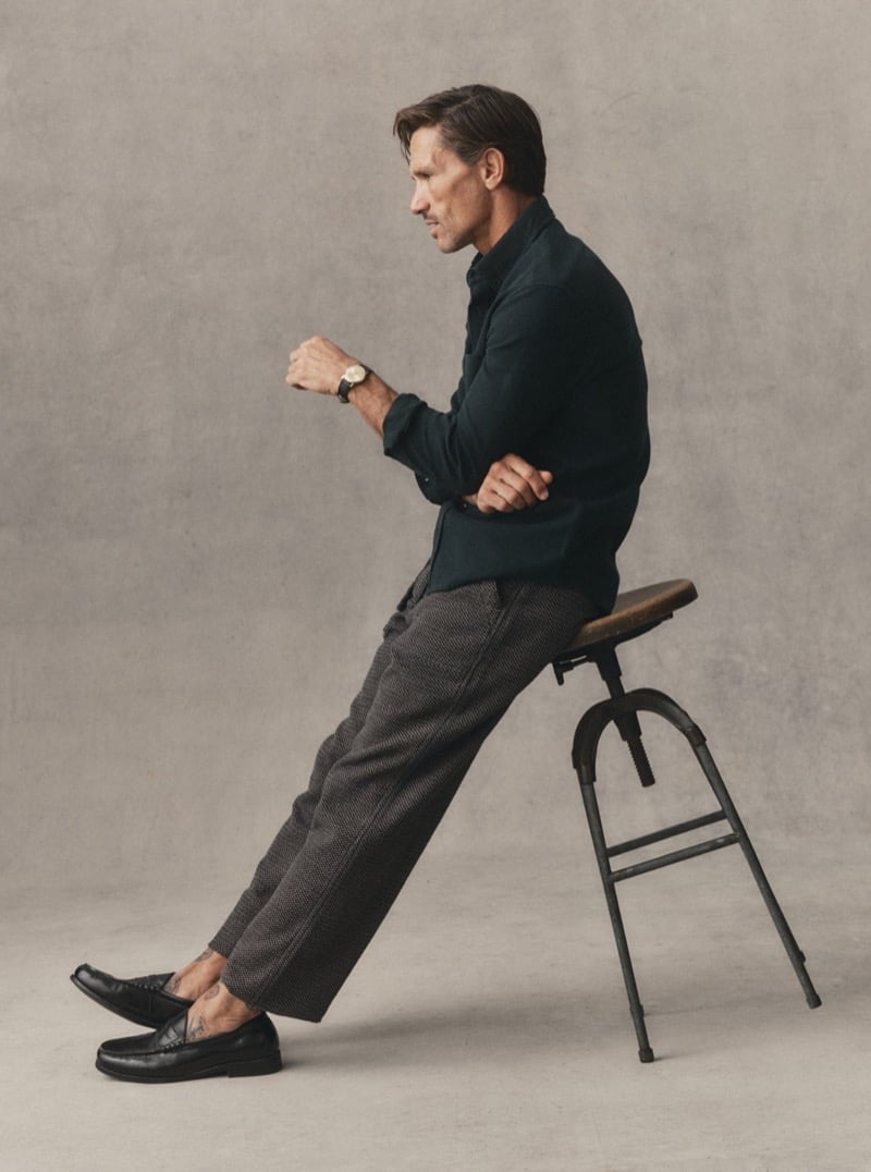 Man in a dark green shirt and tweed trousers seated on a stool in a studio, for Taylor Stitch Fall 2026