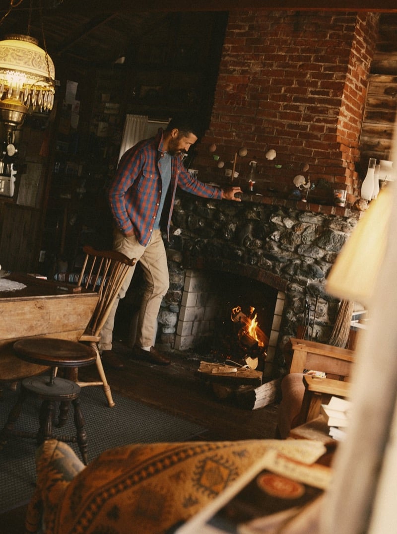 Man in a blue and red plaid flannel shirt standing beside a stone fireplace indoors, for Taylor Stitch Fall 2026