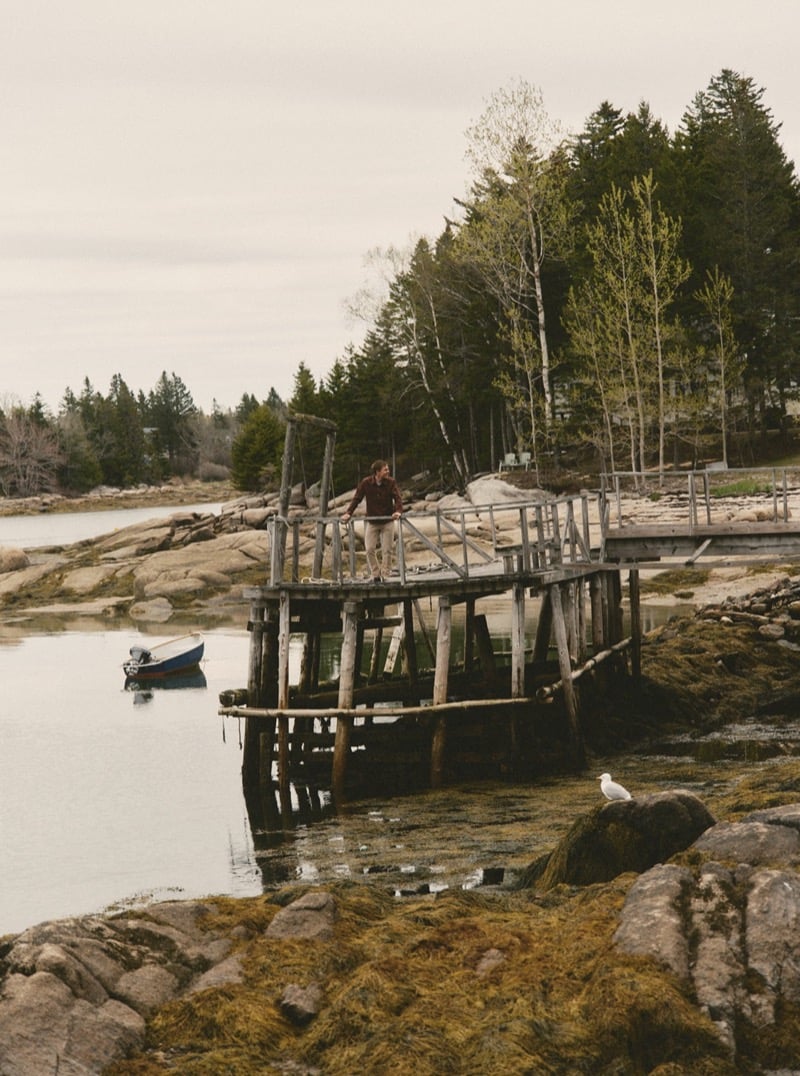 Man standing on a weathered wooden dock overlooking the water with a small boat moored nearby, for Taylor Stitch Fall 2026