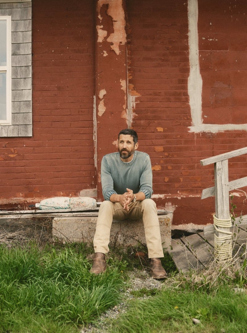 Man in a blue-gray sweater and khaki pants sitting on a stone step against a weathered red building, for Taylor Stitch Fall 2026