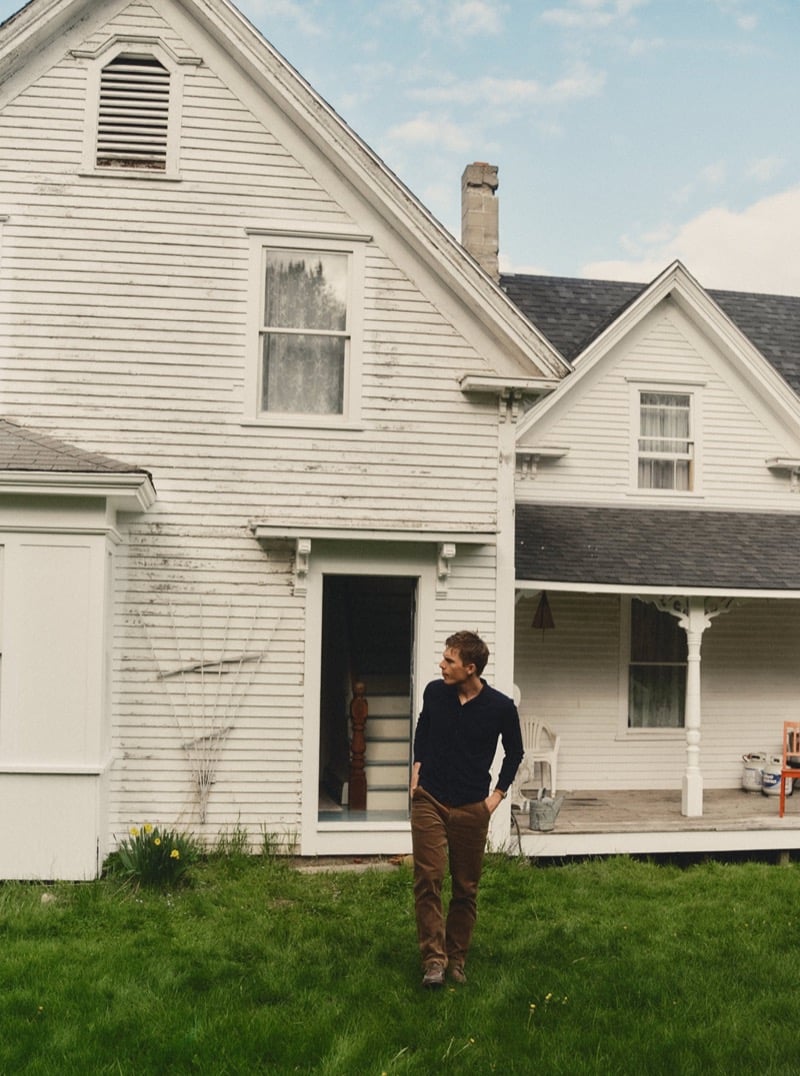 Man in a navy henley and brown pants walking in front of a weathered white farmhouse, for Taylor Stitch Fall 2026