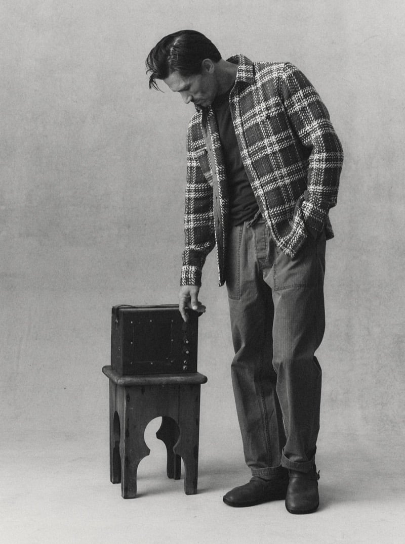 Black-and-white image of a man in a plaid overshirt and corduroy pants reaching for an item on a small wooden table, for Taylor Stitch Fall 2026