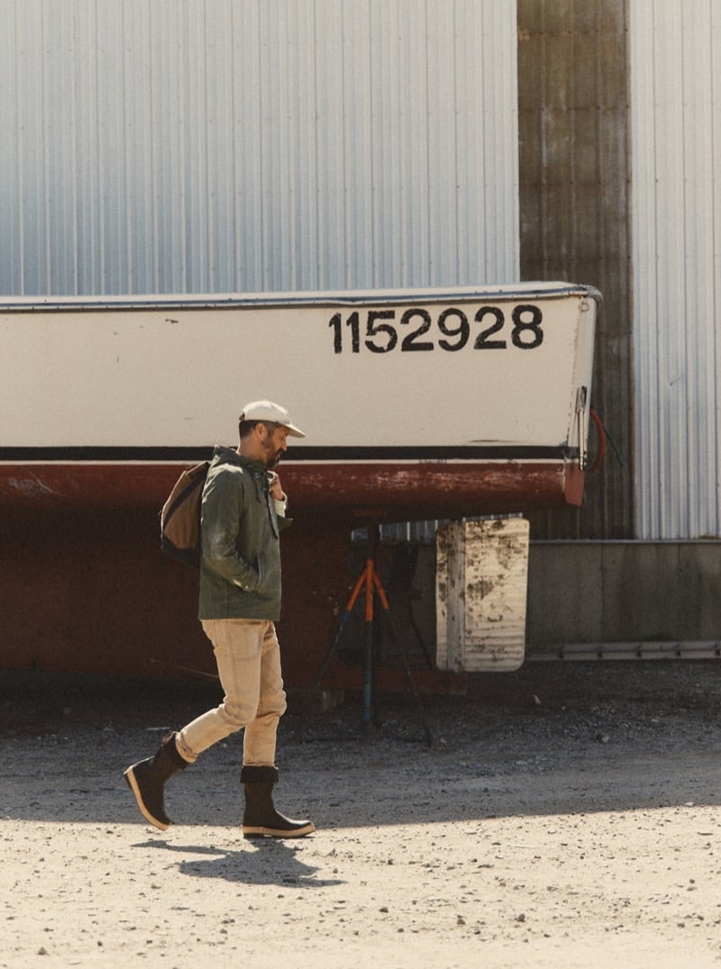 Man in an olive hooded jacket and khaki pants walking past a boat hull, for Taylor Stitch Fall 2026