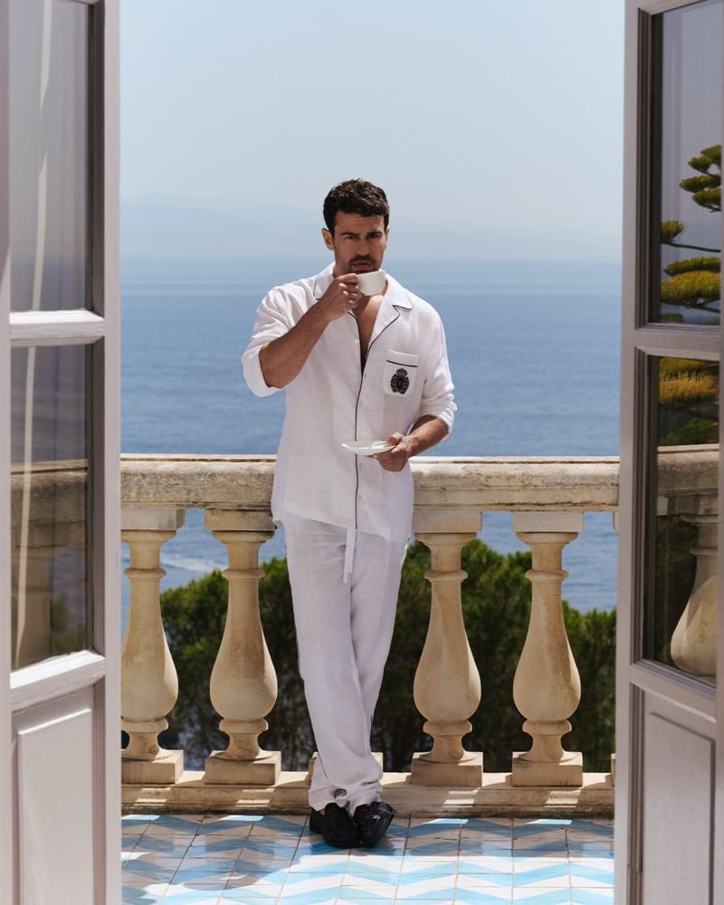 Theo James stands on a blue and white tiled balcony floor in a white linen pajama set, coffee cup raised, with a stone balustrade and the Taormina coastline behind him for Dolce & Gabbana Light Blue Pour Homme.