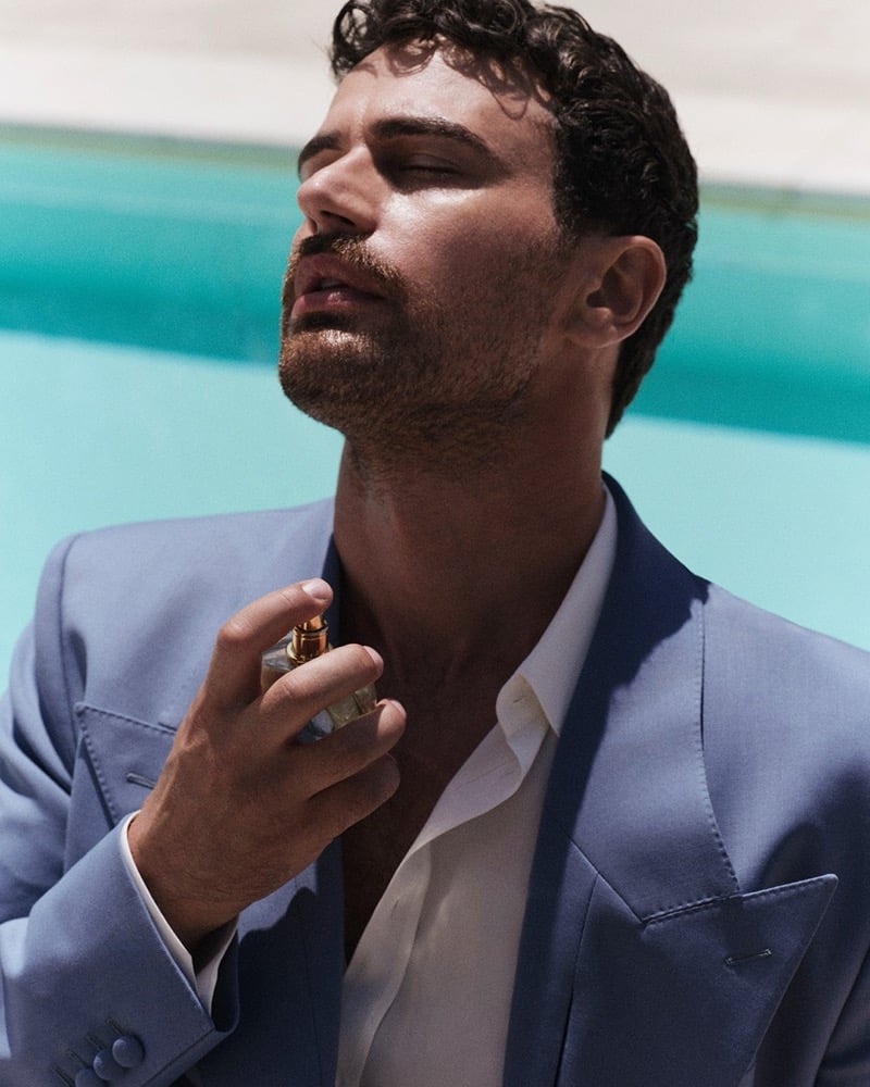 Theo James wears a blue tailored blazer over a white shirt applying Dolce & Gabbana Light Blue Pour Homme to his neck beside a pool in Taormina.