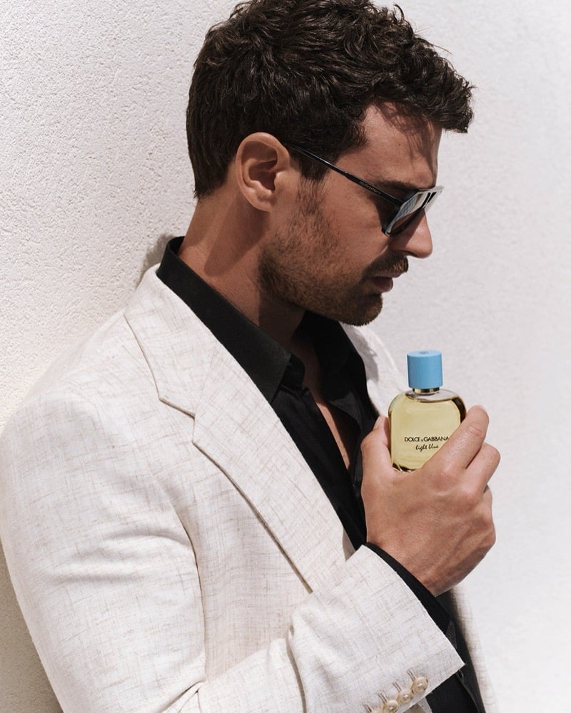 Theo James wears a cream linen blazer over a black shirt and sunglasses holding a Dolce & Gabbana Light Blue Pour Homme bottle against a white stucco wall in Taormina.