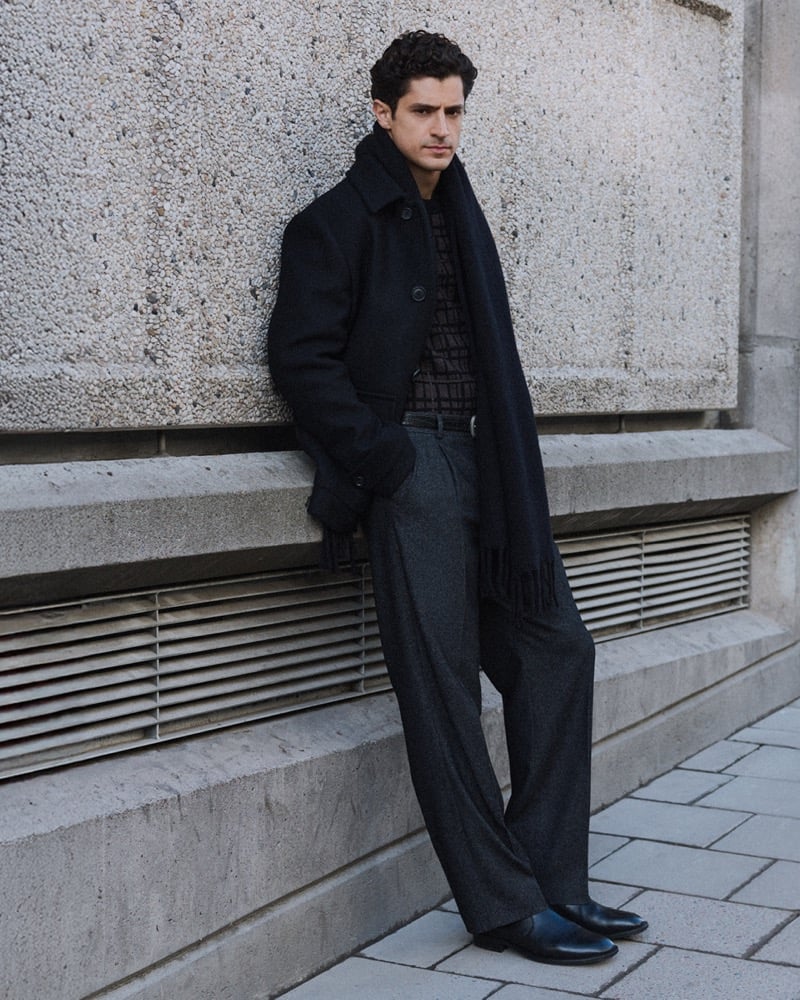 Cyrus Amini leans against a concrete wall in a dark wool coat, checked knit sweater, scarf, and gray wide-leg trousers