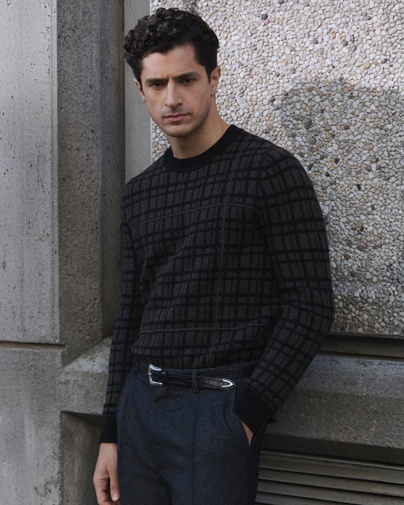 Close-up of Cyrus Amini in a checked knit sweater and dark trousers with a buckled belt, leaning against a textured concrete wall