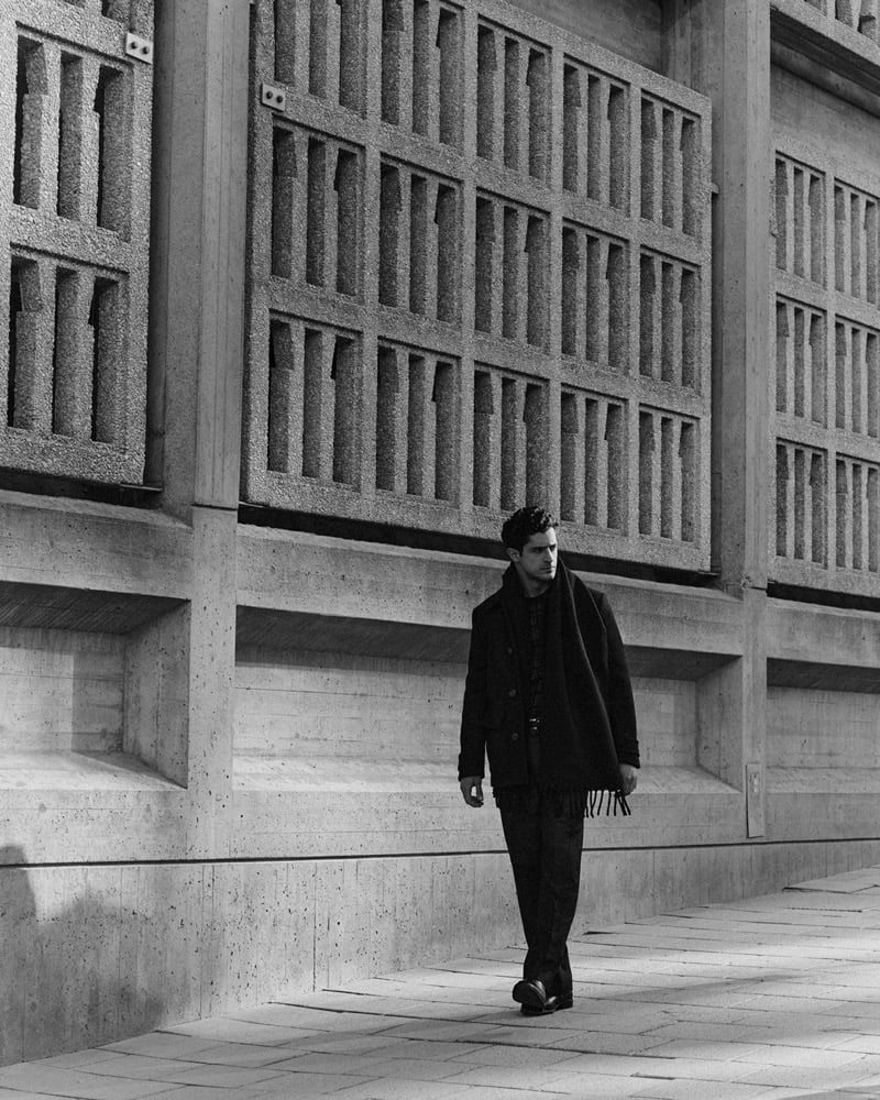 Black-and-white image of Cyrus Amini walking past a brutalist concrete building in a dark coat and scarf
