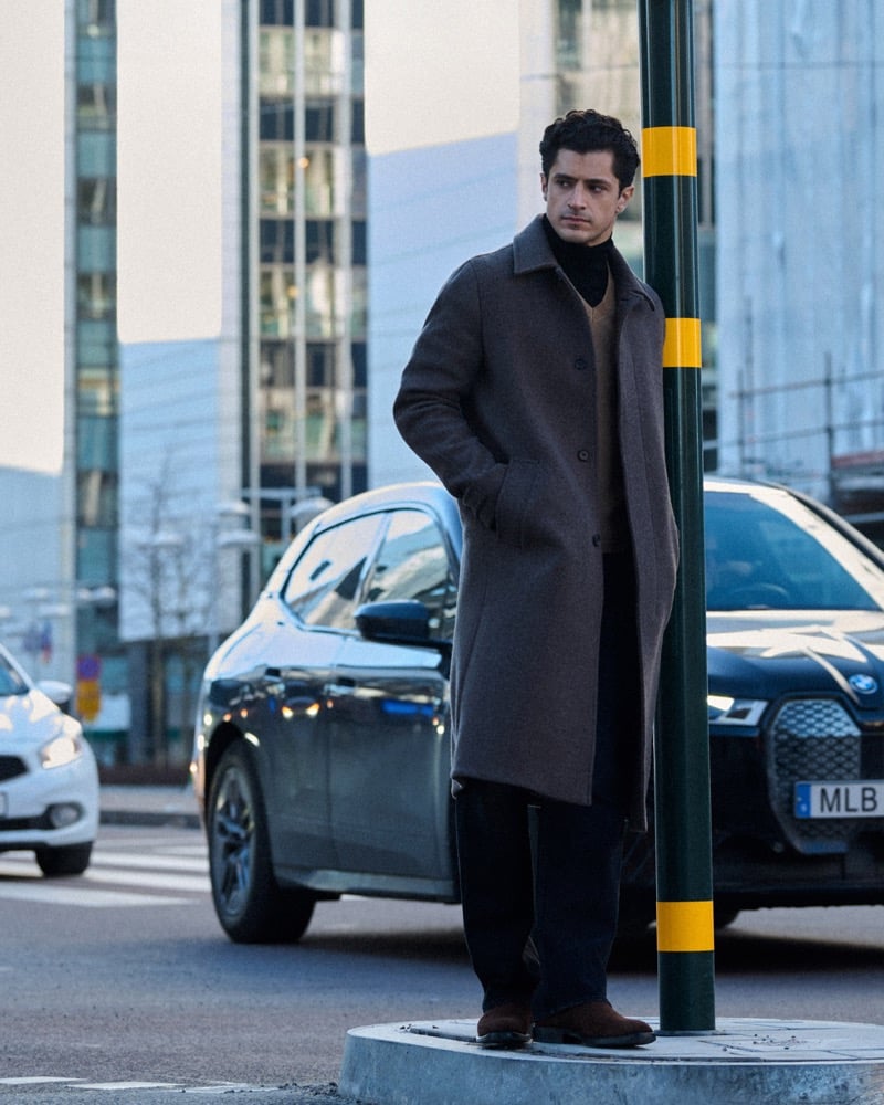 Cyrus Amini stands at a city street corner in a gray wool overcoat over a tan cardigan and dark turtleneck, cars and a lamppost beside him