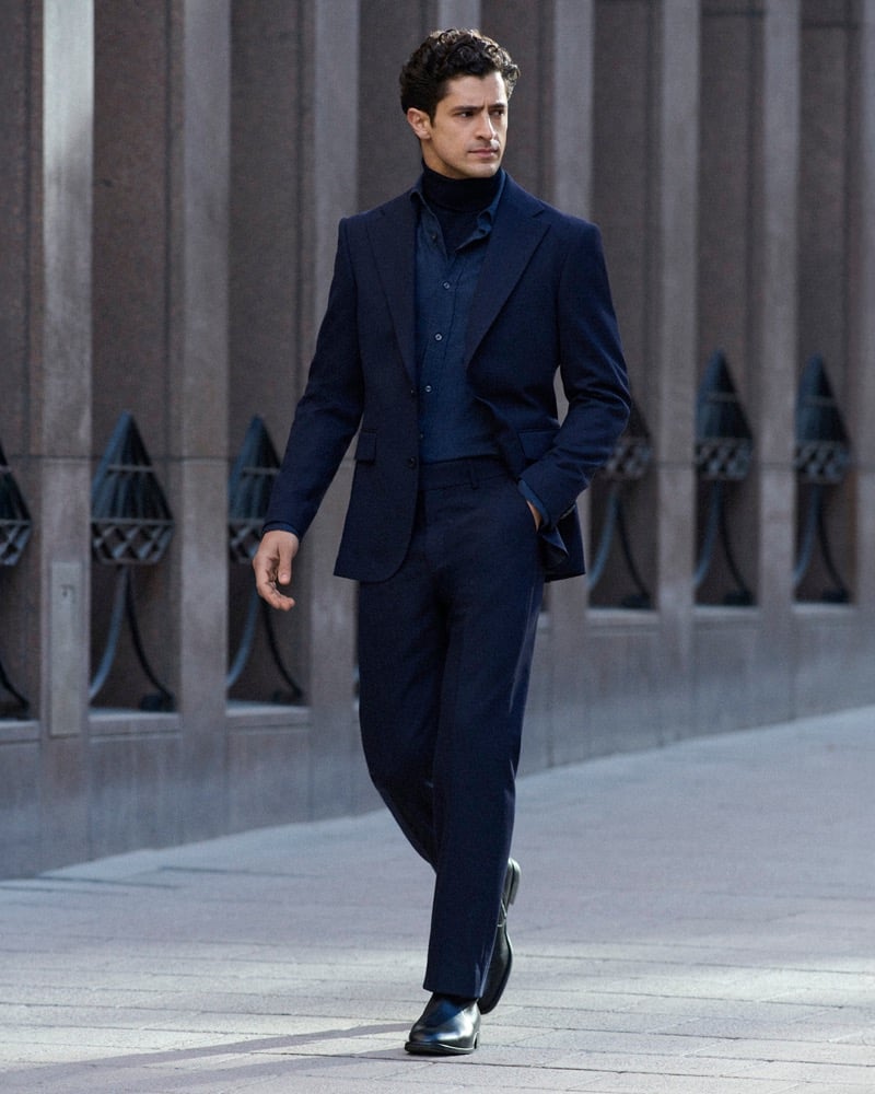 Cyrus Amini walks past a building with lantern-style sconces wearing a navy suit and turtleneck