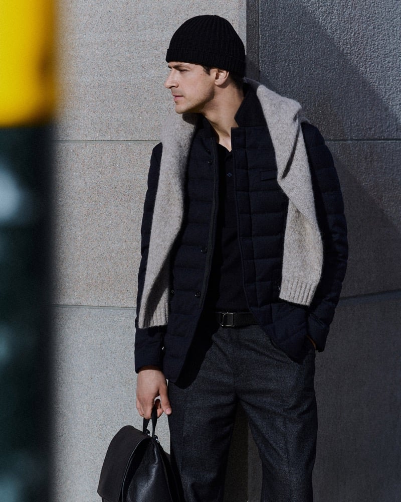 Cyrus Amini leans against a wall in a quilted jacket with a gray knit sweater draped over his shoulders, wearing a beanie and holding a leather bag