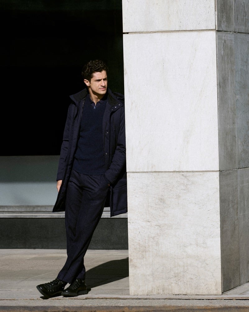 Cyrus Amini leans against a marble column in a hooded coat, dark sweater, and pinstripe trousers