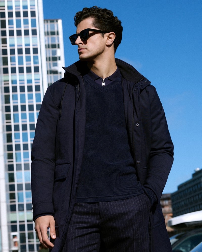 Cyrus Amini wears sunglasses, a hooded coat, and pinstripe trousers, standing before glass towers and a clear blue sky