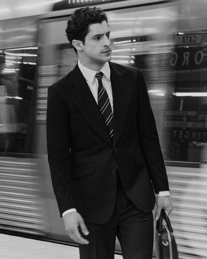 Black-and-white image of Cyrus Amini in a dark suit, tie, and light-colored shirt holding a briefcase as a train blurs past on a subway platform