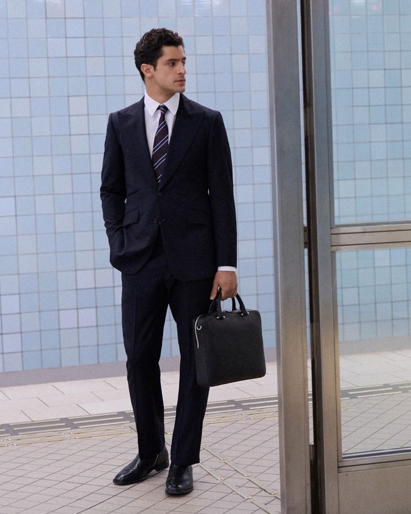 Cyrus Amini stands in a navy suit with a striped tie, holding a black bag, in front of a blue tiled wall