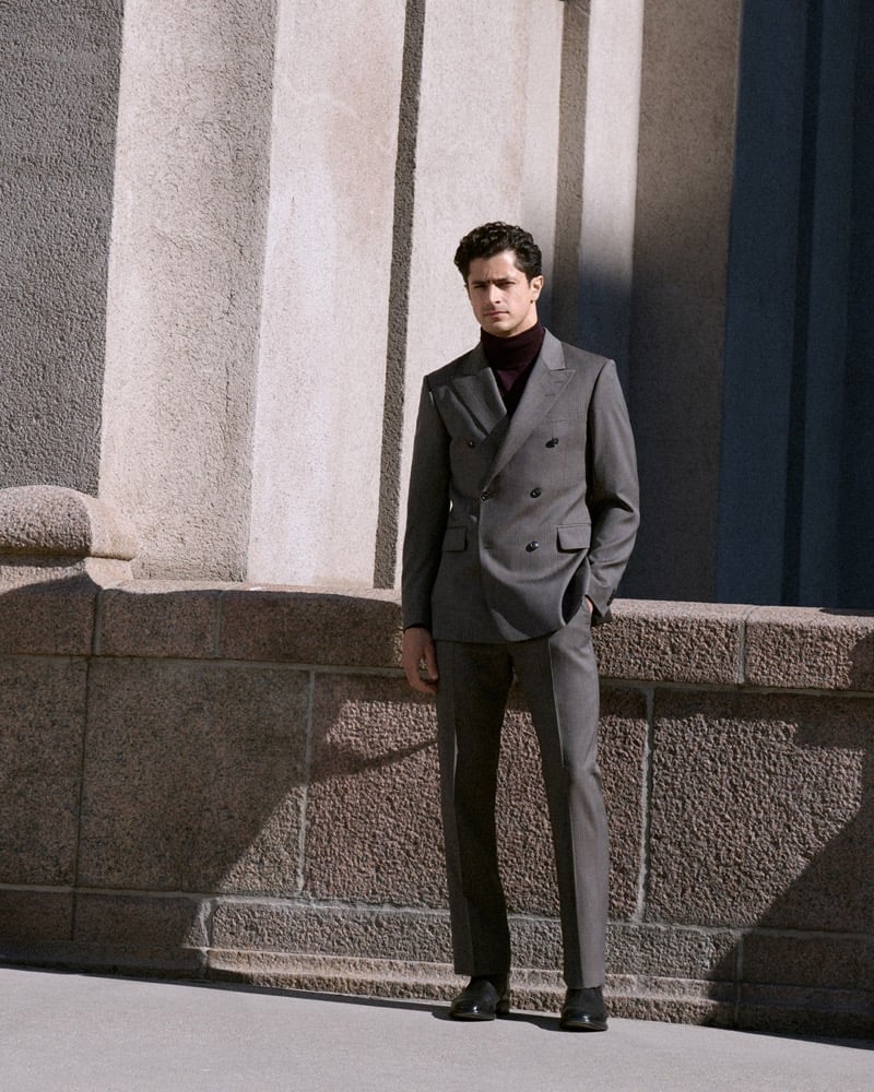 Cyrus Amini stands against a stone building in a gray double-breasted suit and a burgundy turtleneck
