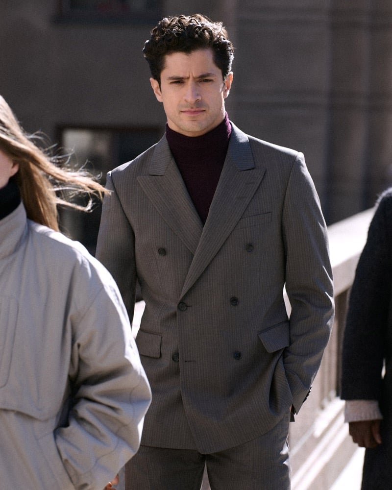 Cyrus Amini stands on a bridge among other pedestrians in a gray double-breasted suit and a burgundy turtleneck