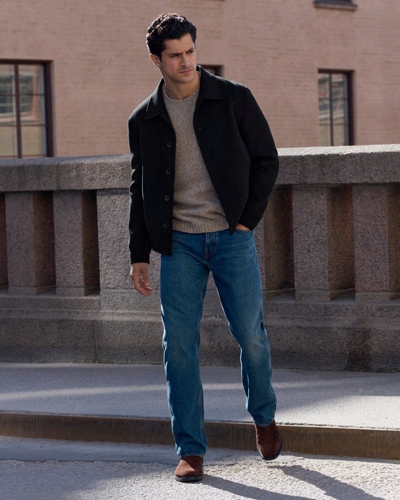 Cyrus Amini walks across a stone bridge in a black wool jacket, gray sweater, and blue jeans