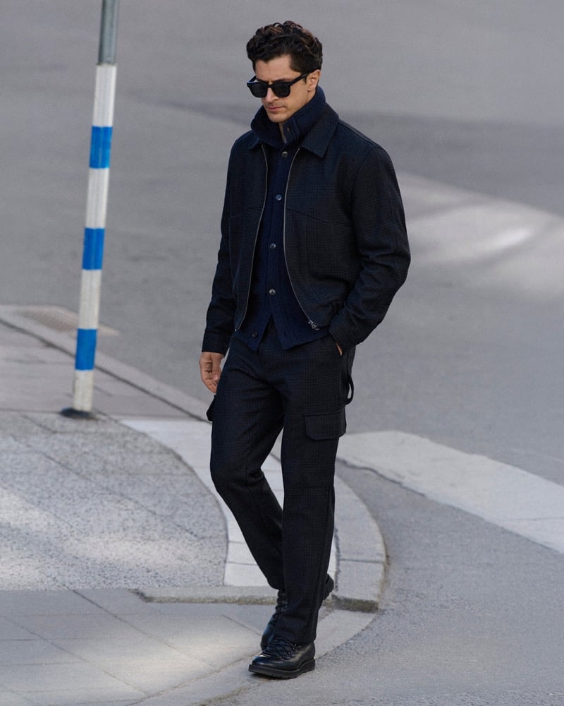 Cyrus Amini wears sunglasses and a checked wool jacket over a zip sweater while crossing a street beside a striped pole