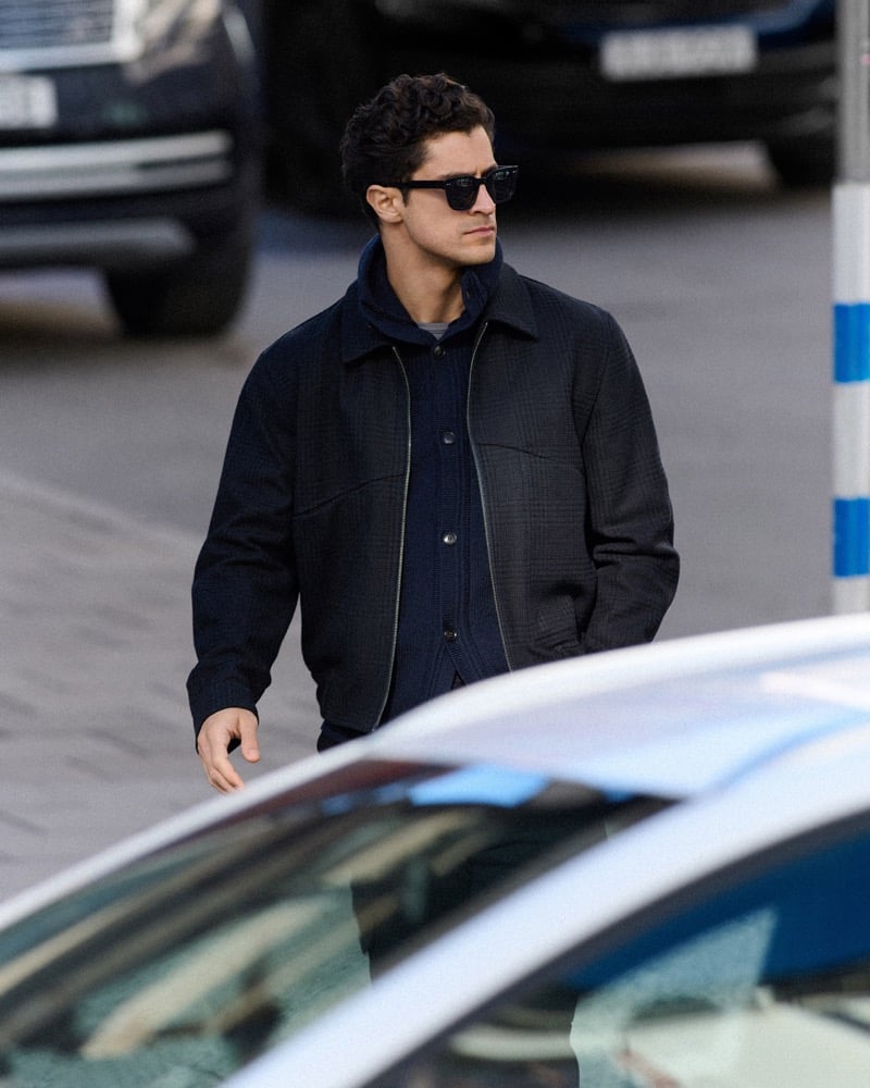 Close-up of Cyrus Amini in sunglasses and a checked wool jacket over a zip sweater, cars parked behind him