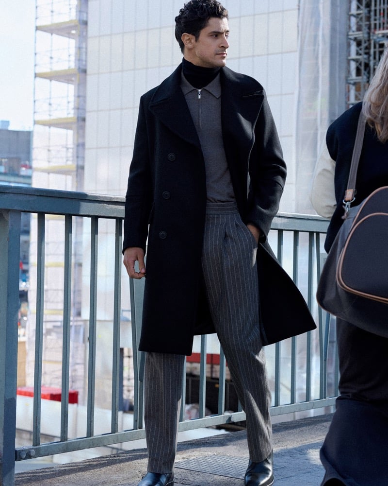 Cyrus Amini stands on a walkway in a black double-breasted overcoat, gray zip turtleneck, and pinstripe trousers, scaffolding visible behind him