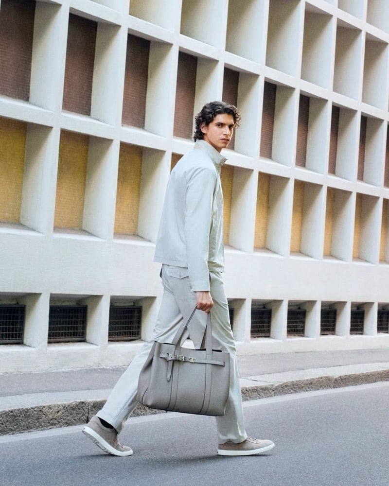 Hedi Ben Tekaya walks alongside a ribbed concrete building in a stone-colored jacket and trousers, holding a stone tote bag with a belt-buckle strap detail