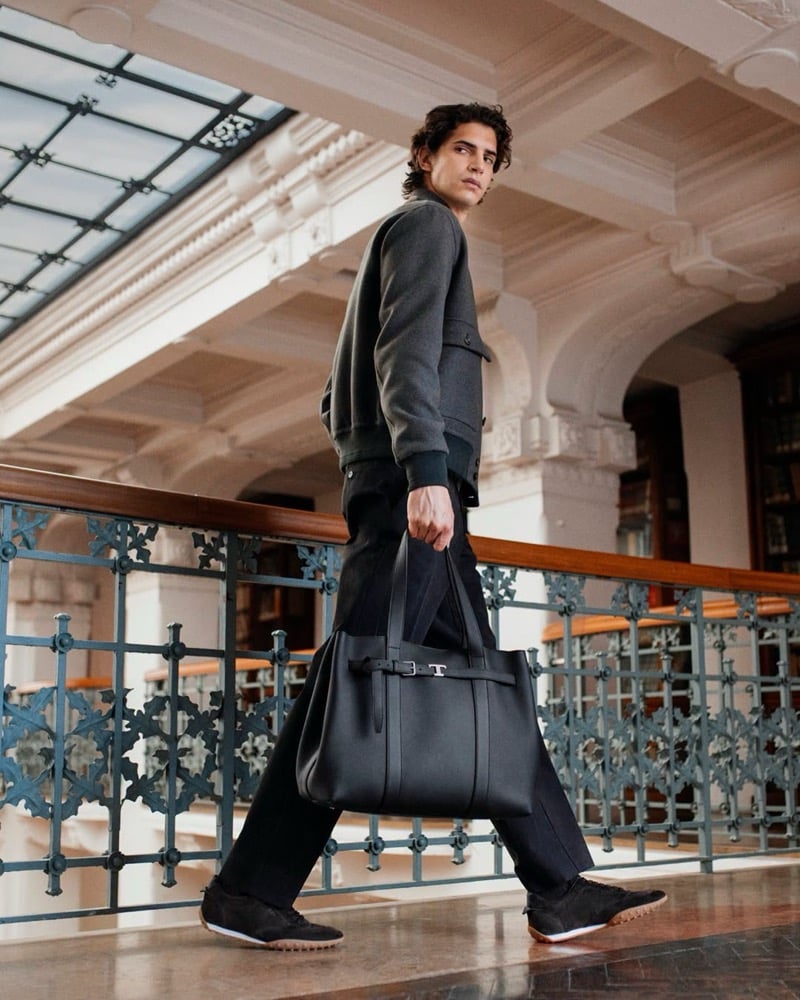 Hedi Ben Tekaya walks along an iron railing under an ornate glass ceiling in a charcoal jacket and dark trousers, holding a leather tote bag
